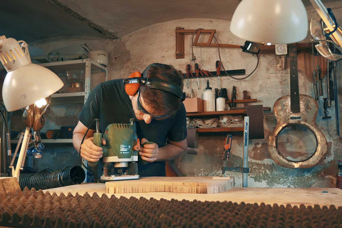 Muž pracuje s horní frézkou na kusu dřeva v dílně. Má ochranné brýle a chrániče sluchu.