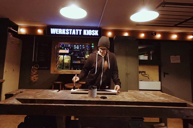 Osoba natírá dřevěný rám na pracovní lavici s nápisem Werkstatt Kiosk v pozadí.