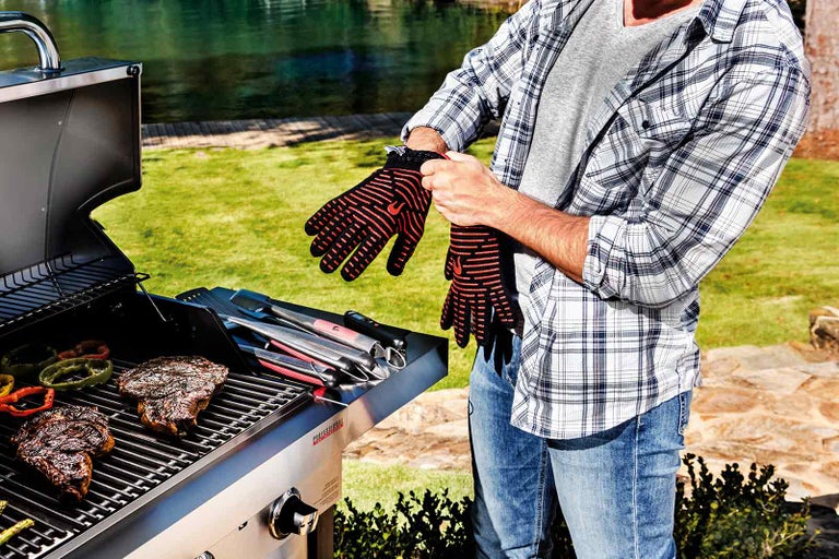 Muž si navléká grilovací rukavice pro ochranu před teplem, zatímco se na grilu připravují steaky a zelenina.