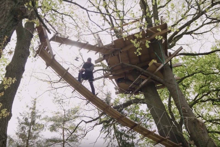 Muž balancuje na dřevěném mostě k domku na stromě