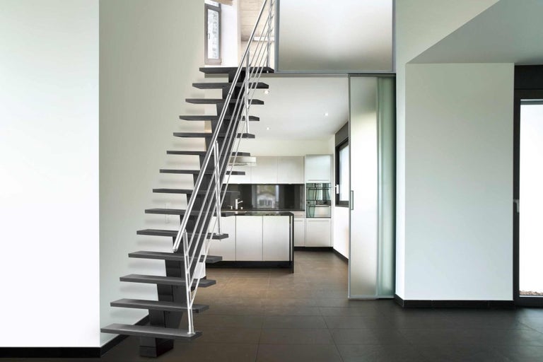 Interiérový snímek moderního domu s samonosným dřevěným schodištěm a nerezovým zábradlím, které vede do kuchyně.