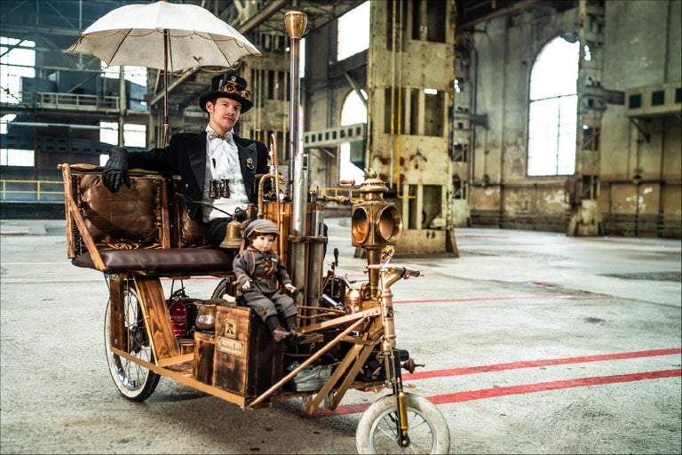 Muž ve stylu steampunk na zdobené tříkolce s panenkou v průmyslové hale