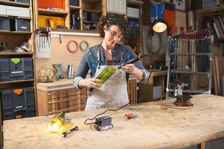 Žena vrtá otvor do zelené skleněné láhve elektrickým nástrojem v dílně