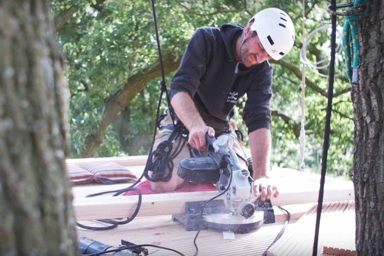 Muž s ochrannou helmou řeže dřevo pokosovou pilou při stavbě domku na stromě.