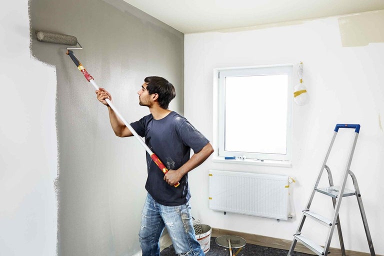 Muž maluje stěnu válečkem na barvu v místnosti.