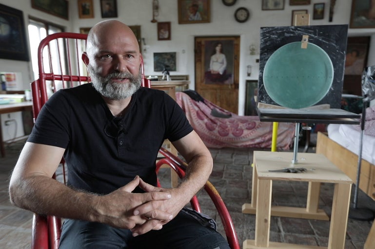 Muž sedí na červené židli před malířským stojanem v světlé místnosti.