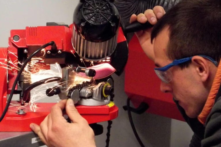 Muž v ochranných brýlích brousí pilový řetěz na elektrické brusce na řetězy, přičemž odlétají jiskry.