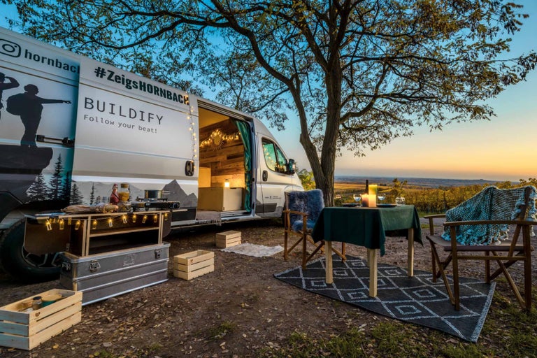 Scéna s přestavěným obytným vozem, stolem, židlemi a venkovním kuchyňským koutem při západu slunce.