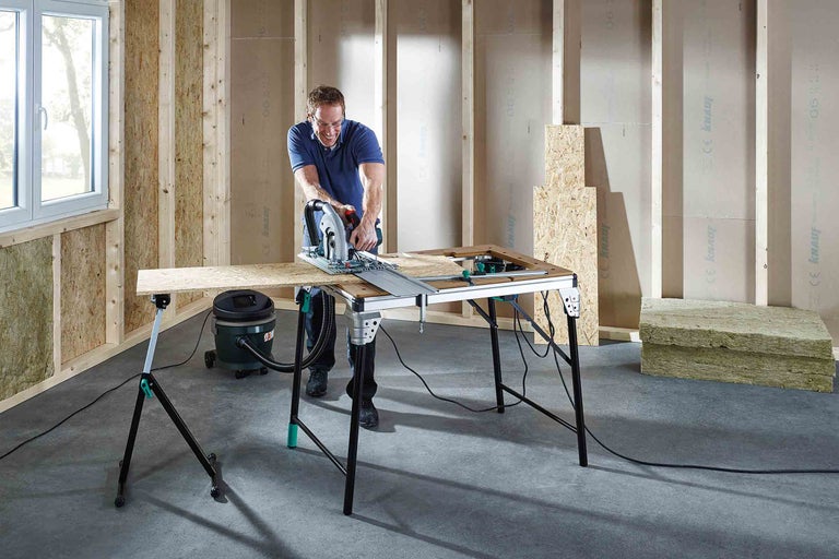 Muž řeže dřevo ruční kotoučovou pilou na pracovním stole v rozestavěné místnosti.