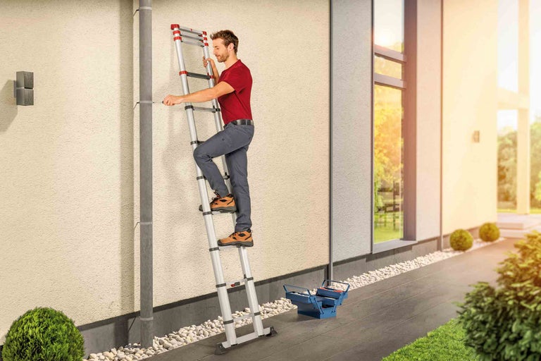 Muž na výsuvném teleskopickém žebříku pracující na okapu, poblíž je skříňka na nářadí