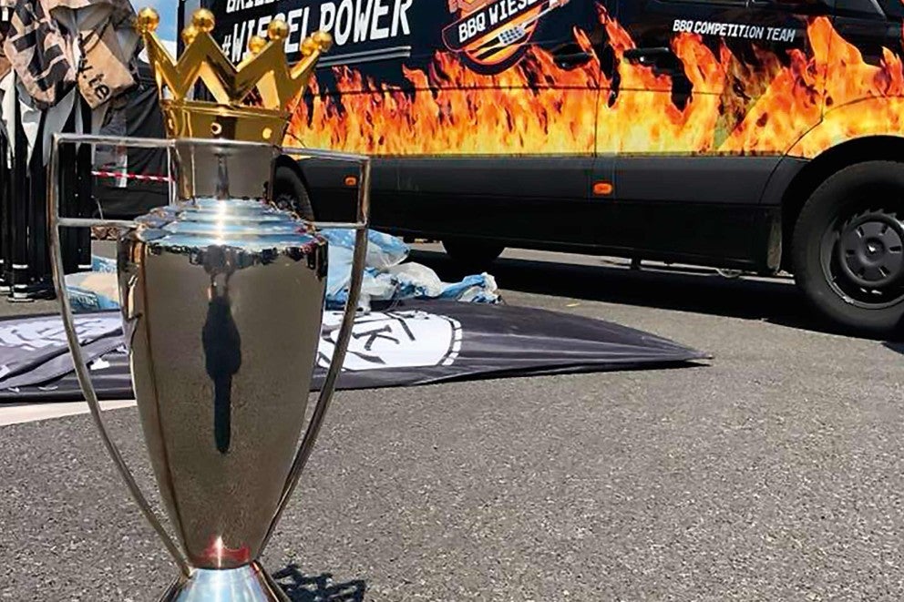 Stříbrná trofej se zlatou korunou před vozidlem s motivy grilu.