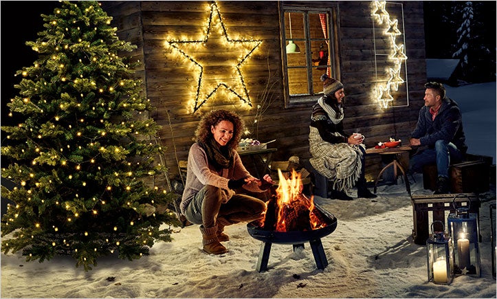 Zimní scéna s lidmi u táboráku, vánoční stromek se světly, hvězdy jako dekorace a dřevěný dům