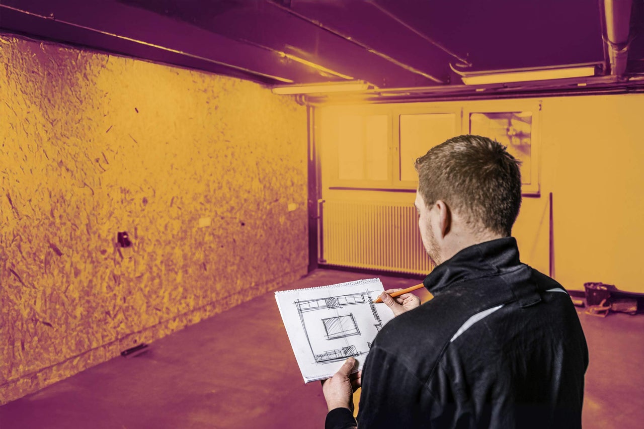 Muž s tužkou a skicákem stojí v místnosti s OSB deskami a plánuje stavební projekt.