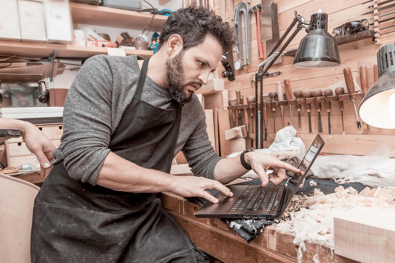 Řemeslník používá notebook ve své dílně, obklopený nástroji a dřevěnými štěpky.