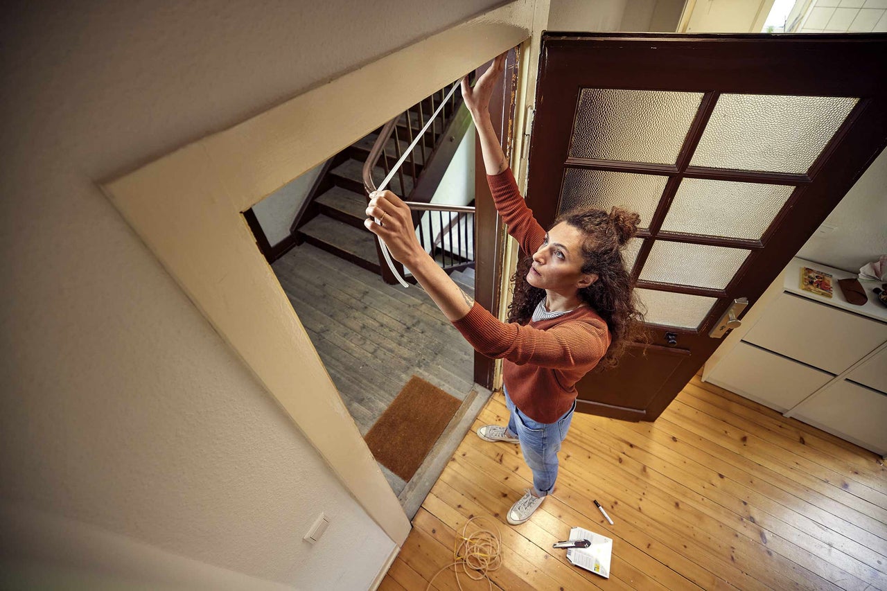 Žena měří těsnicí páskou rám dveří v domě. Na dřevěné podlaze leží nářadí.