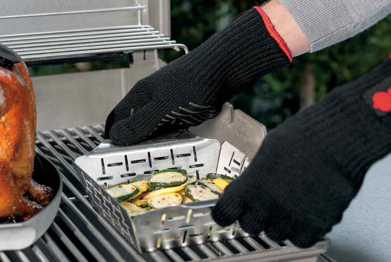 Osoba s grilovacími rukavicemi manipuluje s grilovací mísou plnou grilované zeleniny na grilu