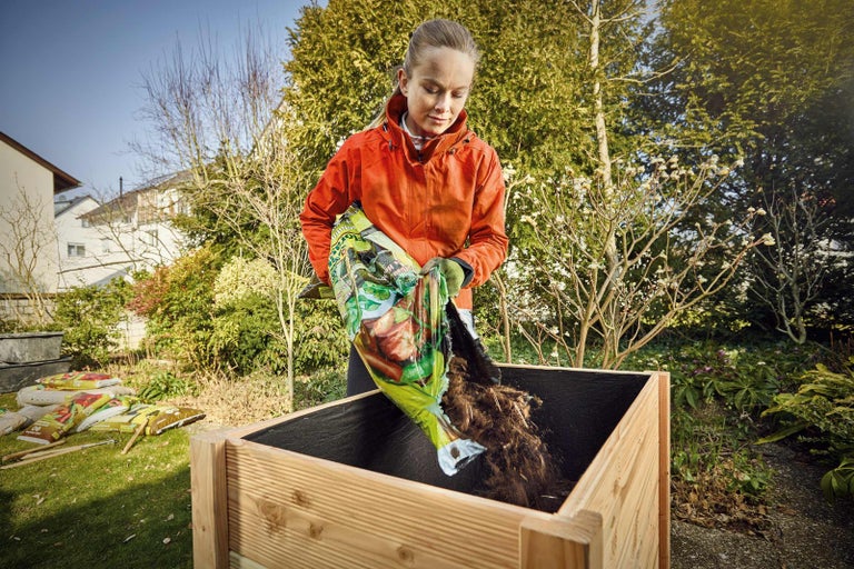 Žena plní vyvýšený záhon zahradní zeminou na zahradě.