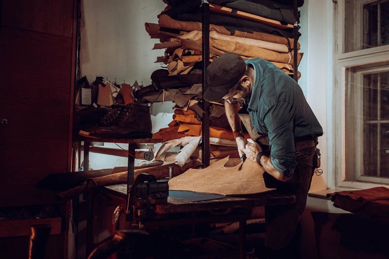Řemeslník řeže kůži v dílně, v pozadí kožené kusy na policích.