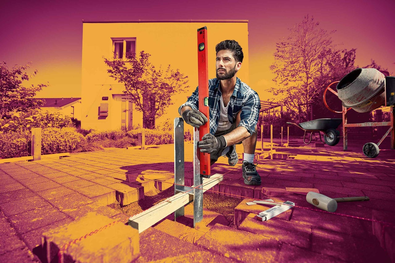 Řemeslník instaluje držák sloupku pomocí vodováhy na zahradě, v pozadí je dům a míchačka na beton.