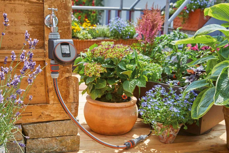 Kohoutek Gardena s ovládáním zavlažování a zavlažovací hadicí v zahradním prostředí s kvetoucími rostlinami.