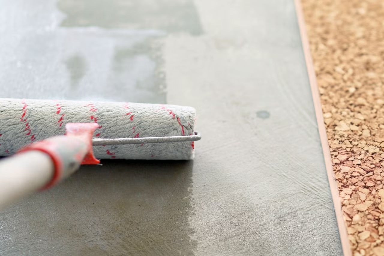 Malířský váleček nanášející penetraci nebo lepidlo na betonový povrch vedle korkové podlahy.