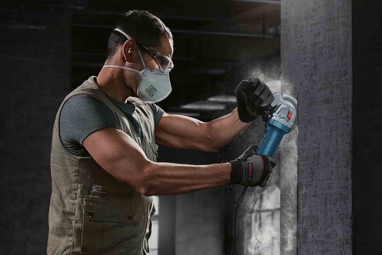 Muž brousí betonovou stěnu úhlovou bruskou, používá osobní ochranné prostředky, jako jsou ochranné brýle, ochrana sluchu, protiprachová maska a rukavice
