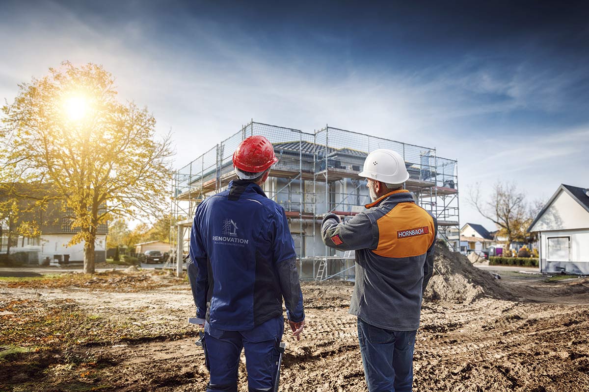 Dva stavební dělníci před rozestavěným domem s lešením. Jeden ze stavebních dělníků má na sobě helmu a pracovní oděv s logem Hornbach.