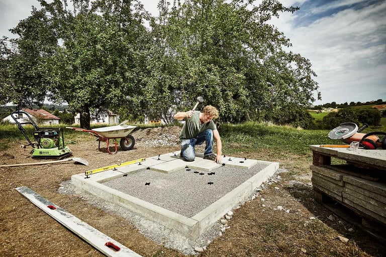 Muž pokládá chodníkové desky na připravený podklad pomocí kladiva a nivelační pomůcky v zahradě.