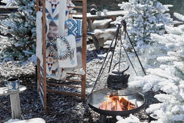 Zimní zahradní scéna s ohništěm, kotlíkem, přikrývkami na žebříku a zasněženými stromy.