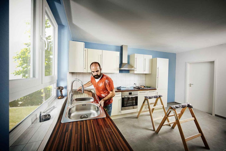 Muž montuje umyvadlo v kuchyni s kuchyňskými skříňkami, oknem a žebříkem