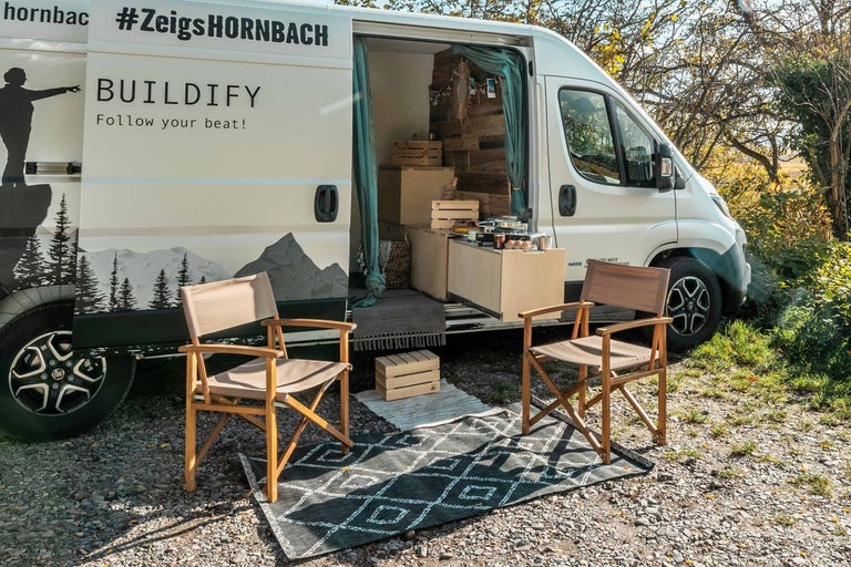 Scéna s přestavěným obytným vozem, dvěma dřevěnými židlemi a venkovním kobercem