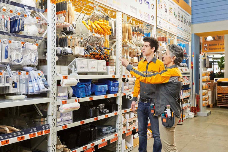 Ve filiálce Hornbach hledají dva zaměstnanci společně produkty v regálech.