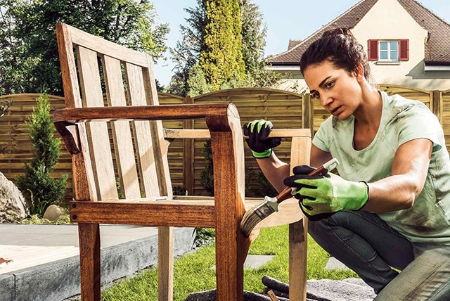 Žena natírá dřevěnou zahradní židli štětcem venku.