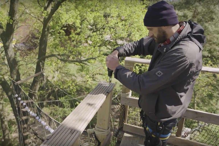 Muž vrtá vrtačkou díru do dřevěné desky na plošině stromu.