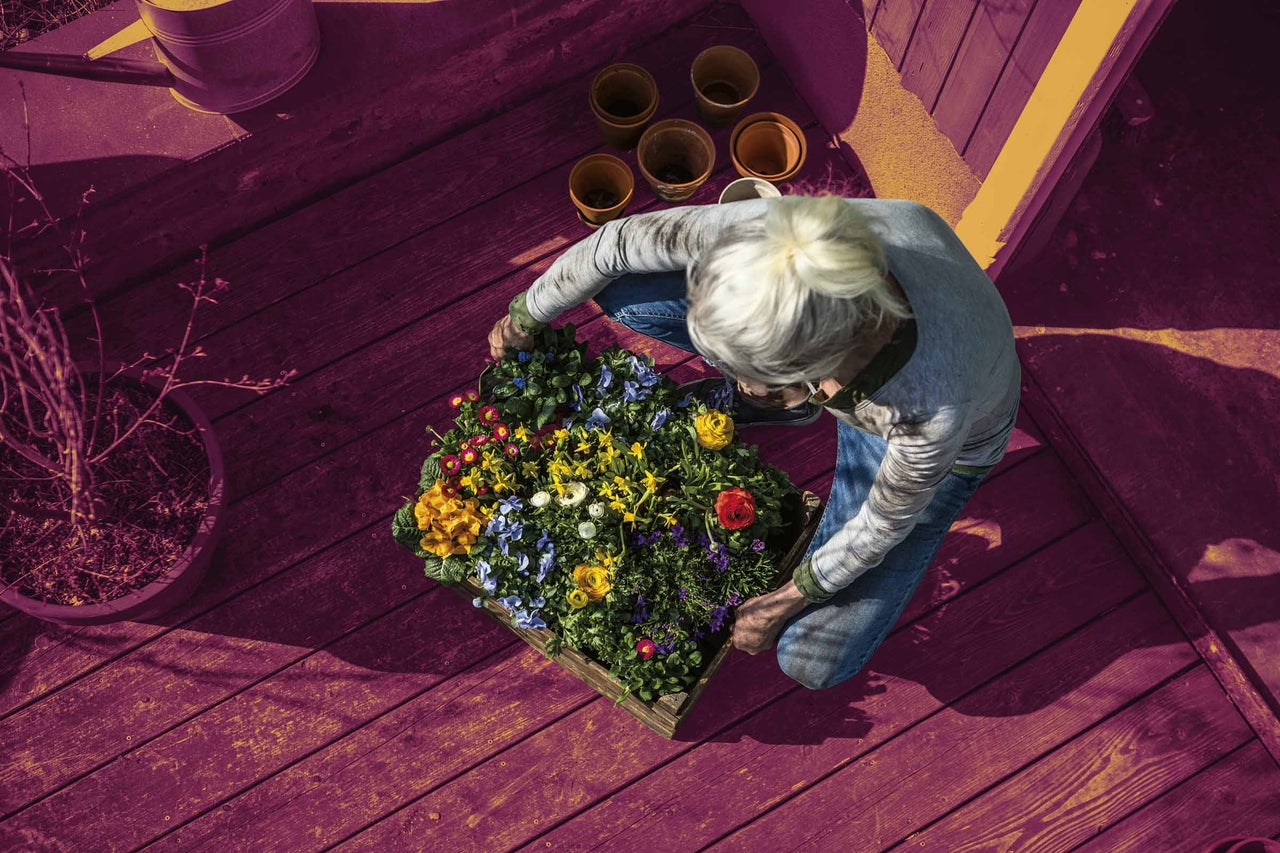 Žena klečí na dřevěné terase a aranžuje květiny do dřevěné bedýnky, kolem níž jsou květináče a konev.