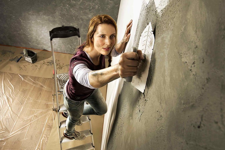 Žena na žebříku nanáší omítku na zeď pomocí hladítka