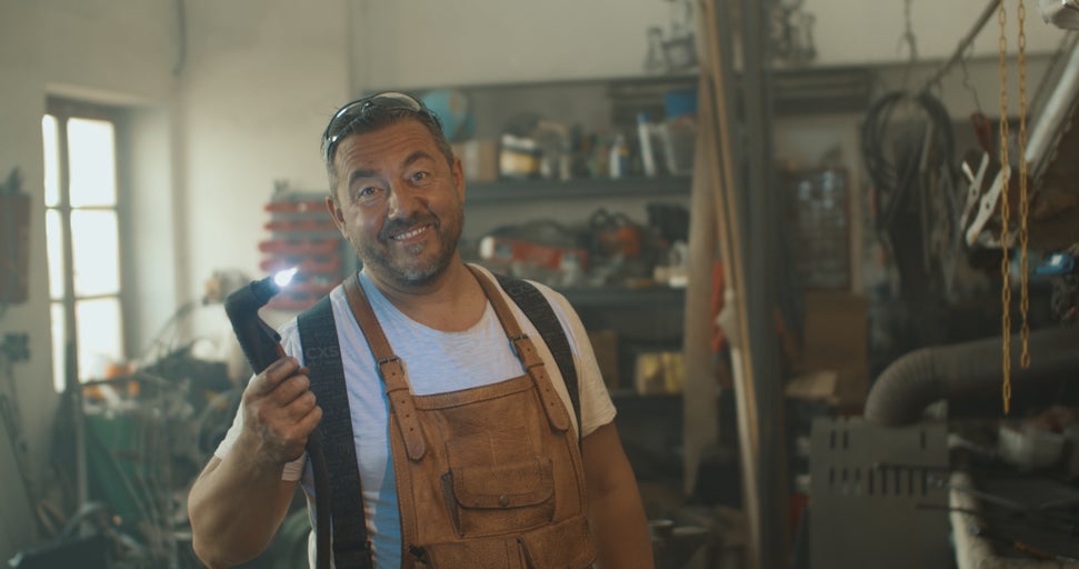 Muž se svářečskými brýlemi a koženou zástěrou drží hořák v dílně