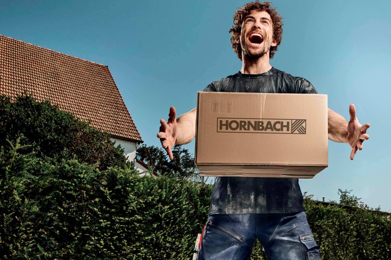 Muž drží krabici s logem Hornbach na pozadí domu a modré oblohy.
