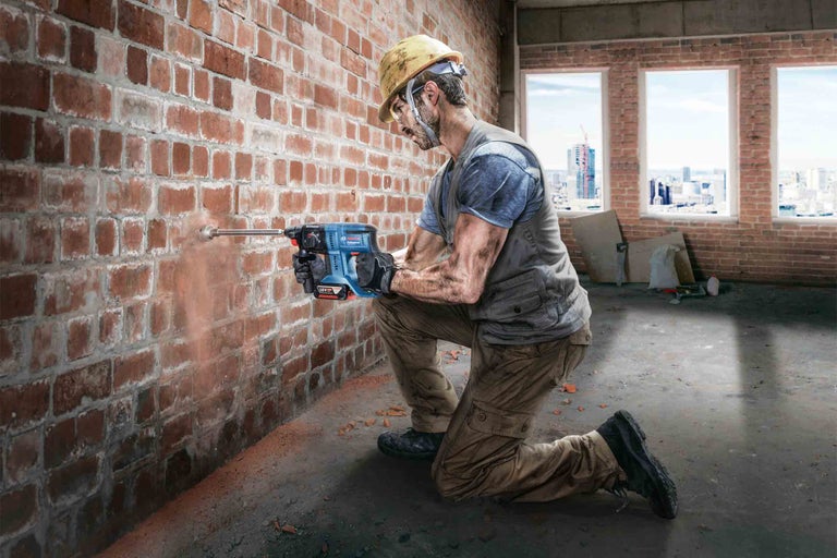 Muž s ochrannou helmou vrtá do cihlové zdi pomocí vrtacího kladiva, v pozadí staveniště.