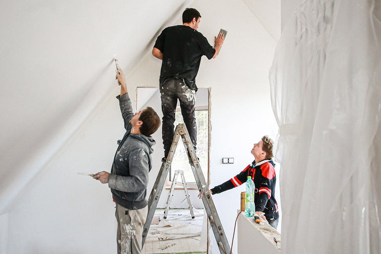 Tři řemeslníci renovují pokoj pomocí žebříků, barvy a brusných nástrojů.