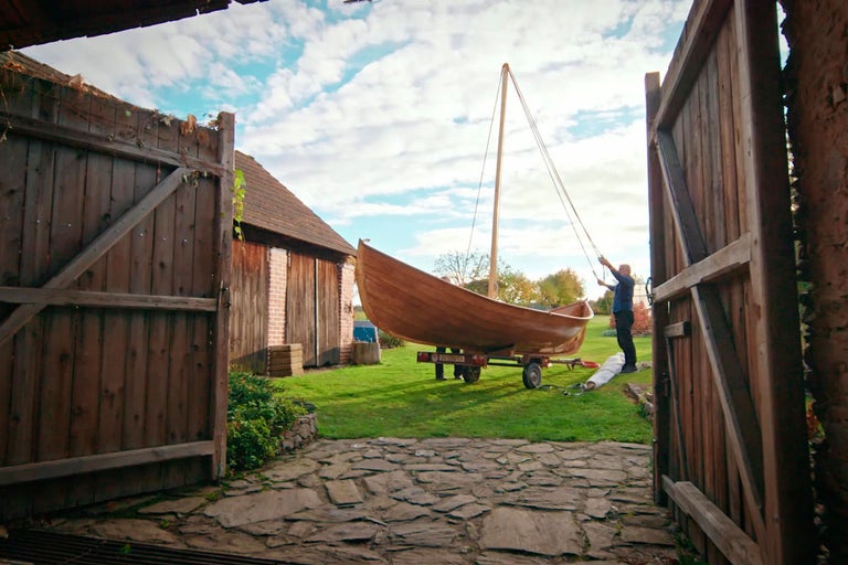 Dřevěná loď se stožárem stojí na přívěsu na louce.