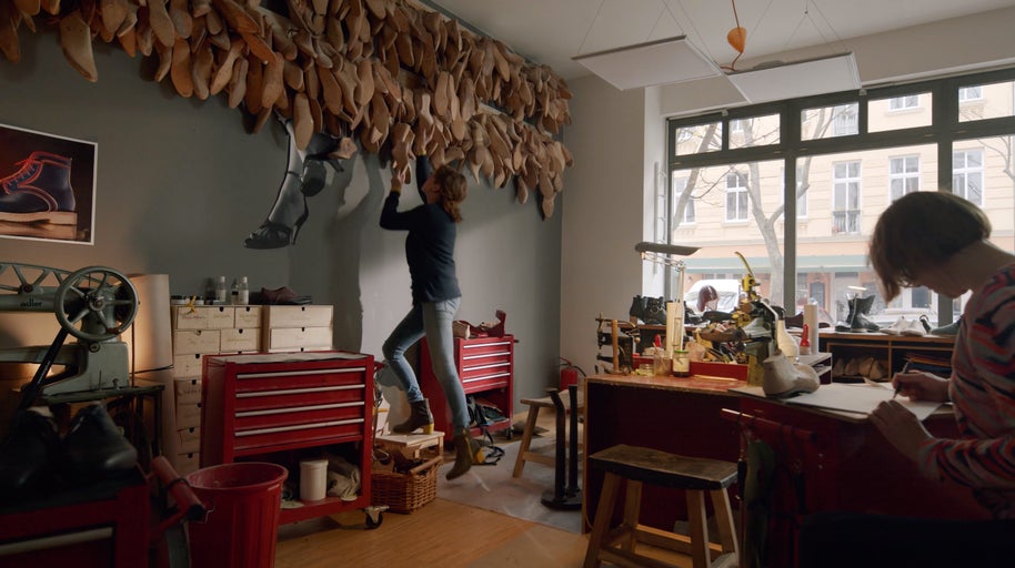 Interiér ševcovské dílny s nástroji, ševcovskými kopyty na zdi a dvěma lidmi při práci