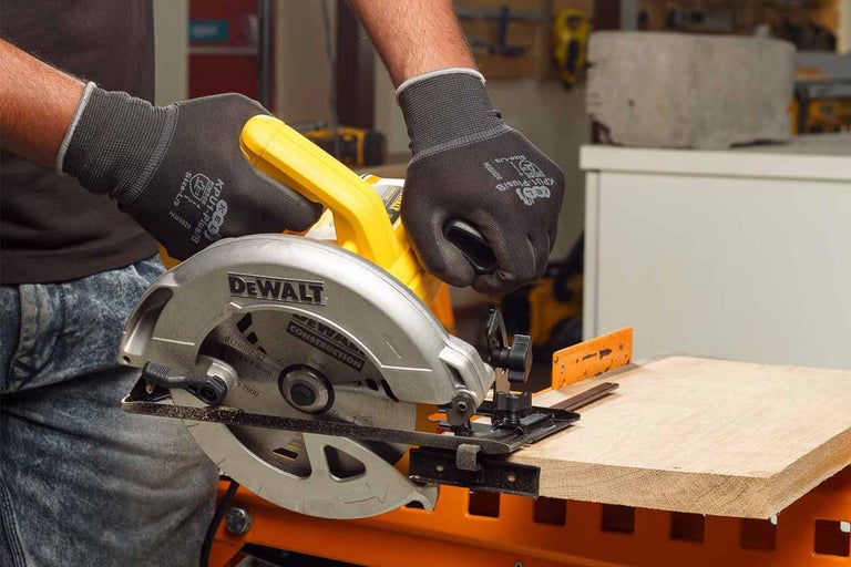 Ruční okružní pila DeWalt řeže dřevěnou desku, přičemž jsou nošeny pracovní rukavice.