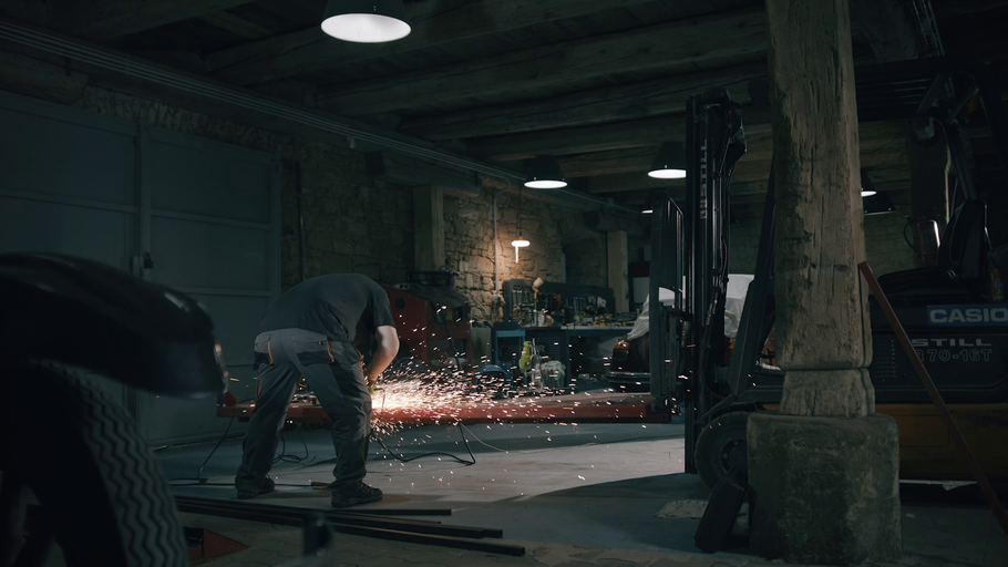 Muž pracuje s úhlovou bruskou v dílně s vysokozdvižným vozíkem v pozadí