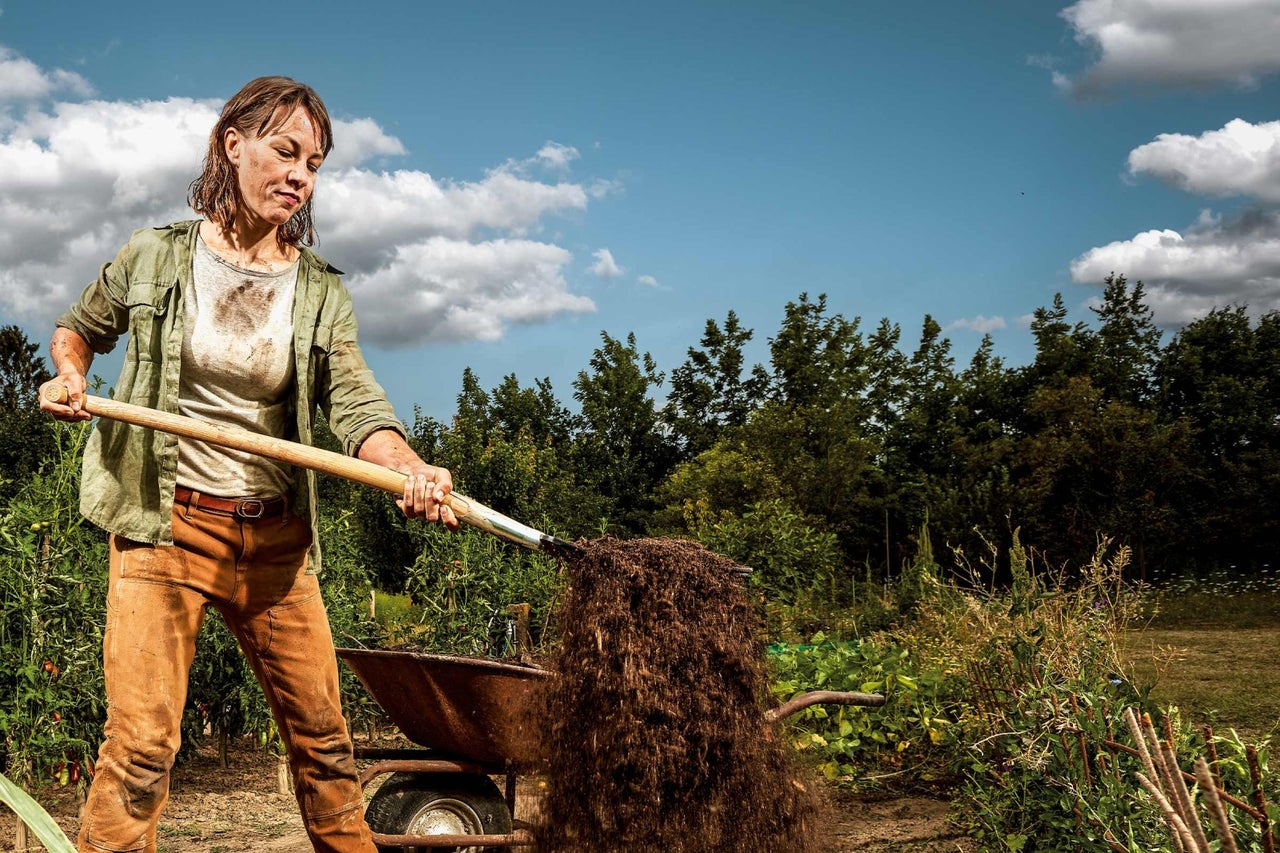 Žena lopatou nakládá hlínu do kolečka na zahradě.