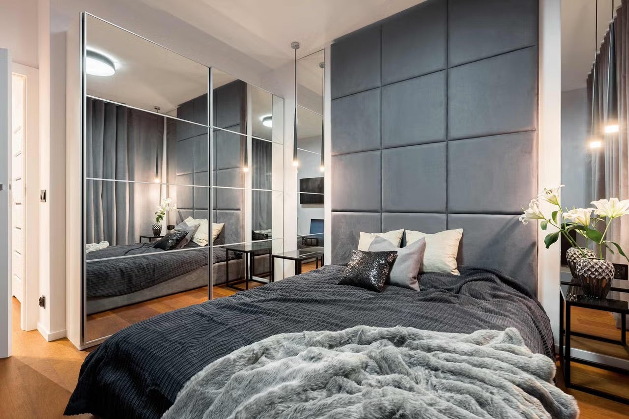 Moderní ložnice s šedými čalouněnými nástěnnými panely, velkou zrcadlovou skříní, postelí a stylovým osvětlením.