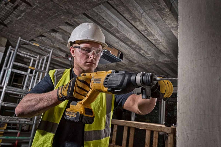 Muž vrtá vrtačkou otvor do zdi. Má na sobě ochrannou helmu, ochranné brýle a ochranné rukavice.
