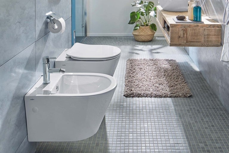 Moderní koupelna se závěsným bidetem a toaletou a šedou koupelnovou předložkou.