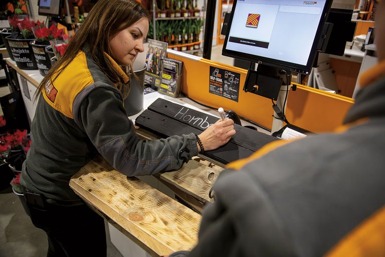 Zaměstnankyně píše na dřevěnou ceduli nápis Hornbach u samoobslužné pokladny