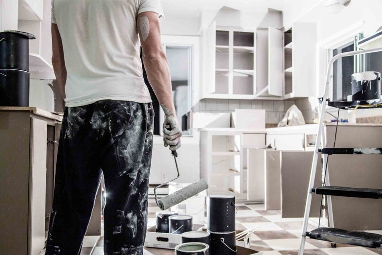 Muž s válečkem na malování ve světlé kuchyni, která se renovuje. Plechovky s barvou a žebřík jsou také viditelné.